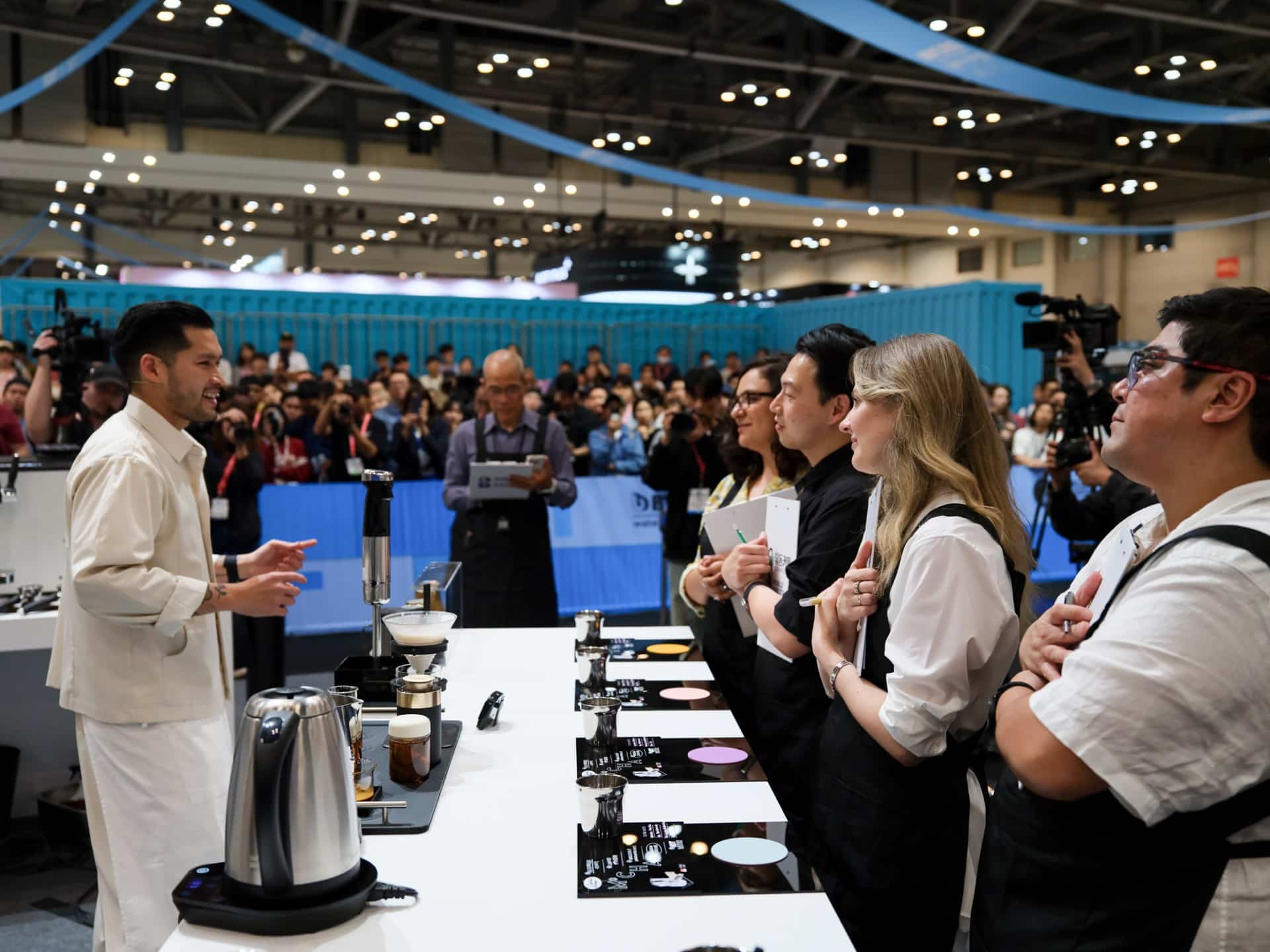 A barista presenting drinks to a row of judges on stage at the World Barista Championship, with a crowd watching behind.